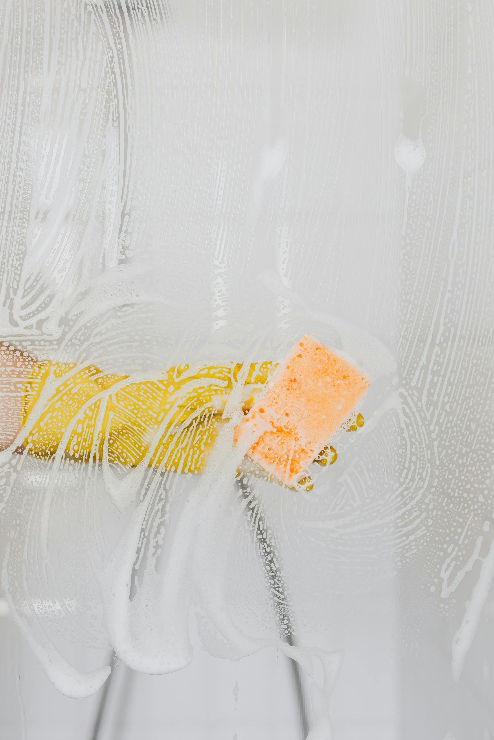 A gloved hand cleans glass with a sponge and soapy water, ensuring hygiene.